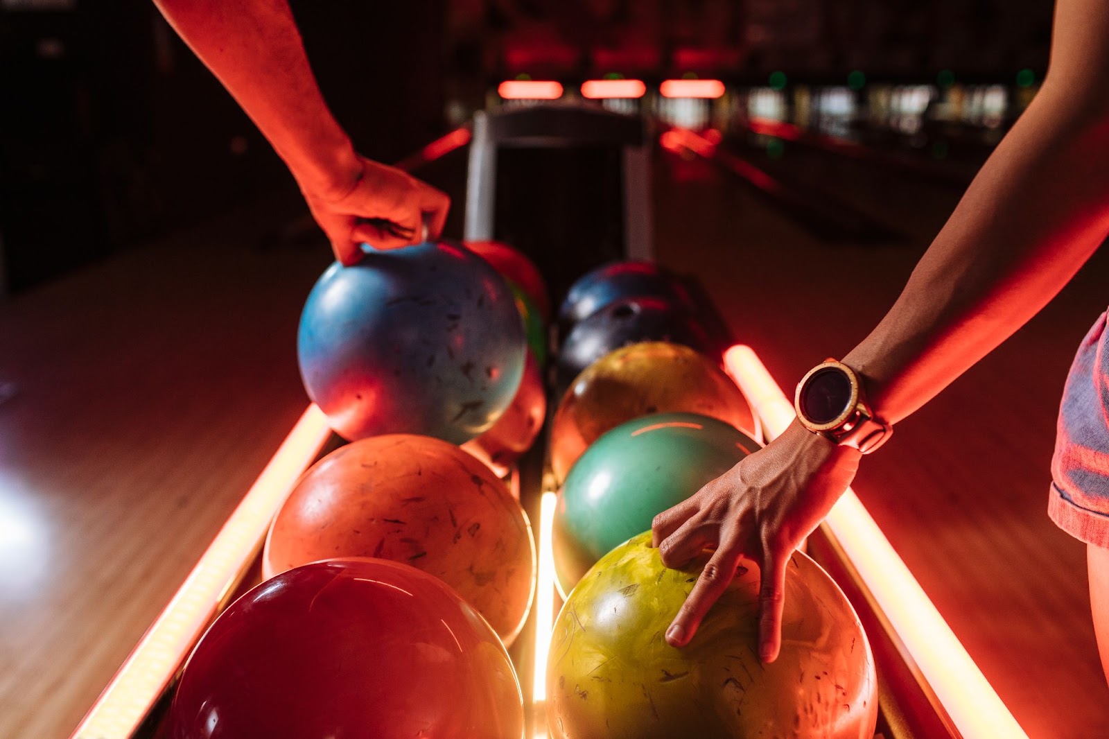 People selecting colorful bowling balls from the rack at a bowling alley under red neon lights.