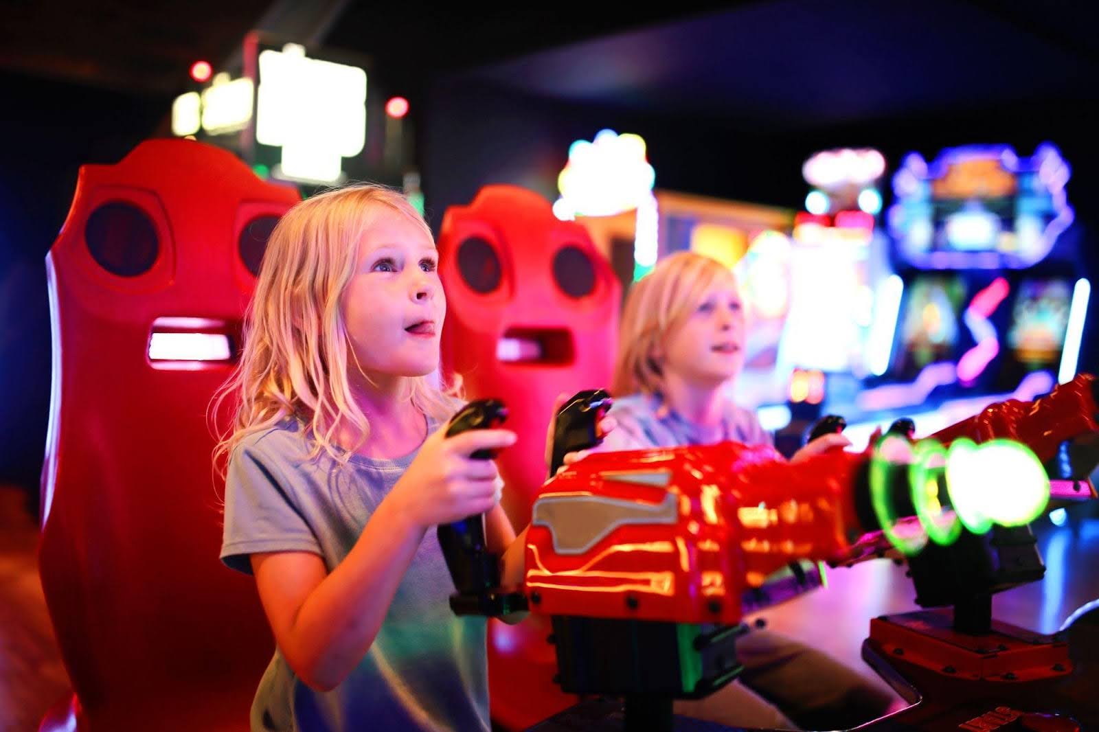 Two young girls joyfully playing video games together in a vibrant arcade filled with colorful machines