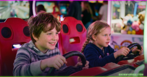 Two kids laughing on racing arcade simulators with pink and green neon glow, indoor entertainment center Ogden Utah