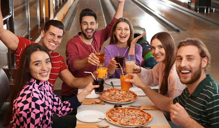 Friends having pizza and drinks at a bowling alley, surrounded by bowling balls and pins.
