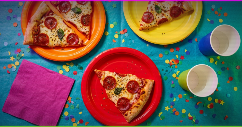 Overhead close-up of pizza slices, paper party plates, cups and scattered confetti on a birthday table